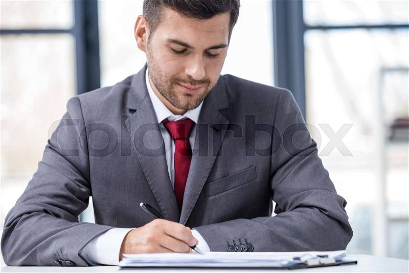 Handsome young businessman sitting at desk and writing in papers, business concept, stock photo