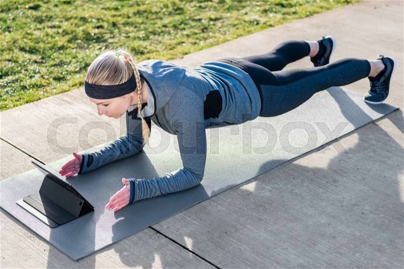 High-angle view of fit young woman watching a motivational video on tablet PC while exercising the plank on mat outdoors in the park, stock photo