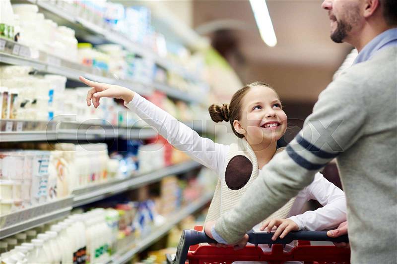 Happy family buying groceries: smiling little girl choosing dairy products from fridge in milk aisle while shopping in supermarket, stock photo