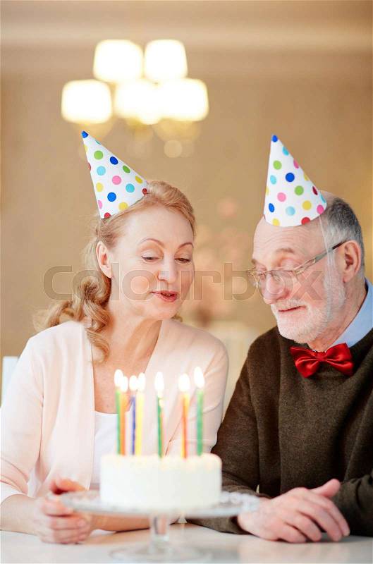 Happy mature man and his wife in birthday caps looking at burning candles in cake, stock photo