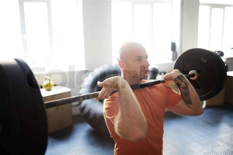 Concentrated bodybuilder doing shoulder press exercise with barbell in gym illuminated with bright sunlight, stock photo