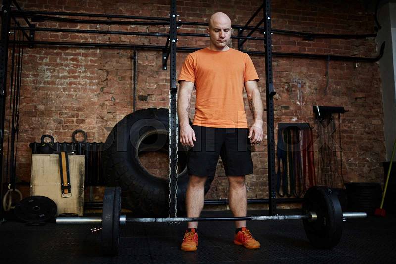 Sporty middle-aged man preparing to do barbell curls, brick wall of spacious gym on background, stock photo