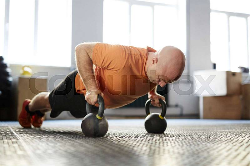 Bald bodybuilder looking down with concentration while doing plank exercise with kettlebells, full-length portrait, stock photo