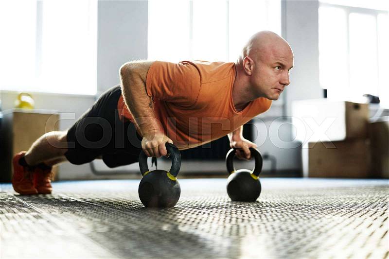 Intensive training in modern gym illuminated with bright sunlight: bald middle-aged sportsman standing in plank position with help of kettlebells, full-length portrait, stock photo