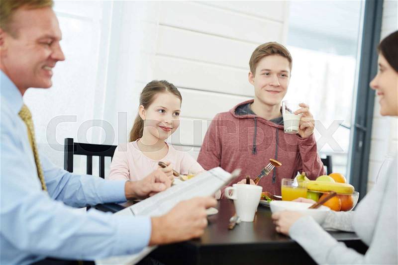 Happy middle-aged parents and their children starting new day with delicious breakfast, handsome father reading newspaper, waist-up portrait, stock photo
