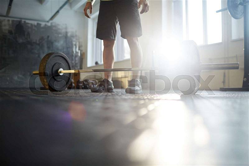 Low section of unrecognizable strong man ready to lift heavy barbell from floor during powerlifting workout in sunlit gym, stock photo