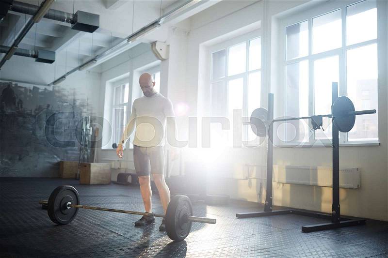 Portrait of strongman ready to lift heavy barbell from floor during workout in sunlit gym, stock photo