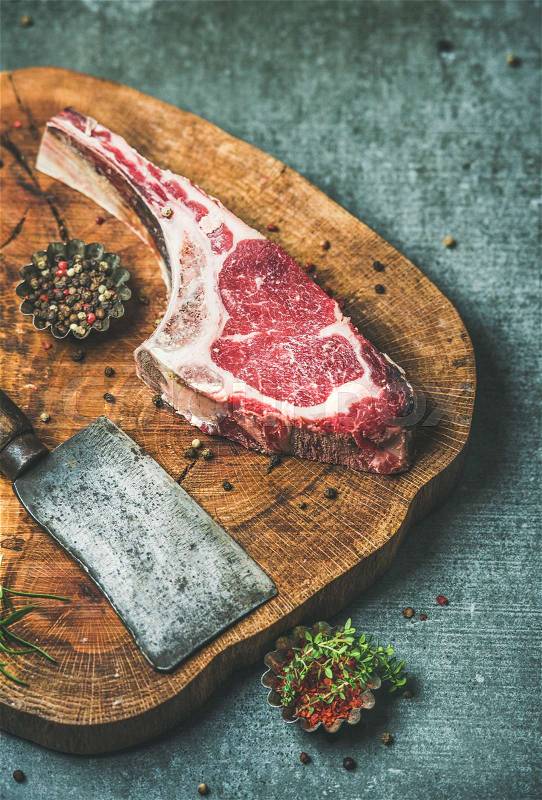 Dry aged raw beef rib eye steak with bone, butcher meat chopping knife and spices on rustic wooden board over grey concrete background, selective focus, stock photo