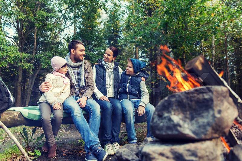 Camping, travel, tourism, hike and people concept - happy family sitting on bench and talking at camp near campfire in woods, stock photo