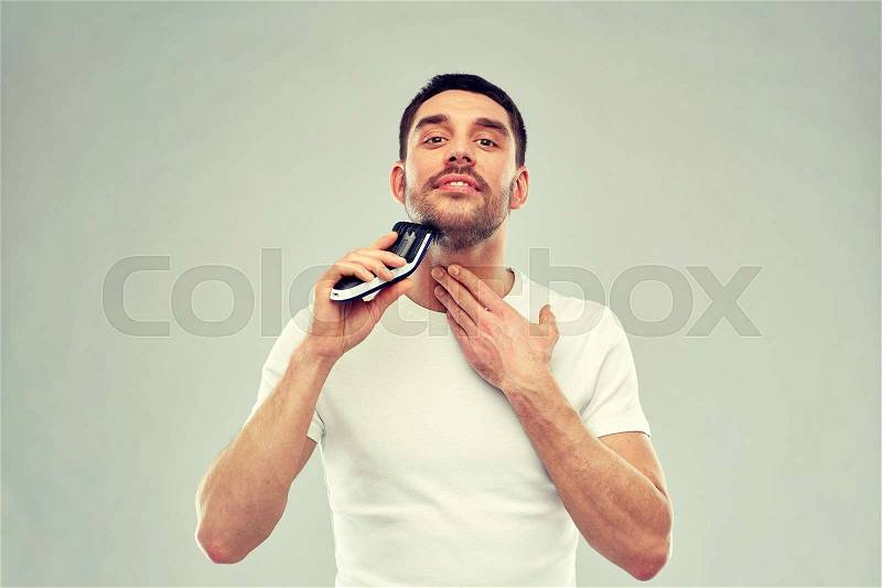 Beauty, grooming and people concept - smiling young man shaving beard with trimmer or electric shaver over gray background, stock photo