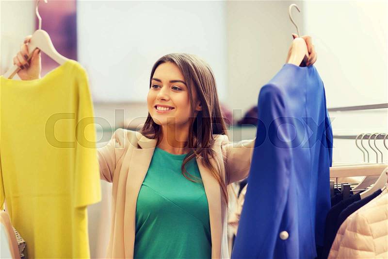 Sale, clothes , shopping, fashion and people concept - happy young woman choosing between shirt and jacket at clothing store, stock photo