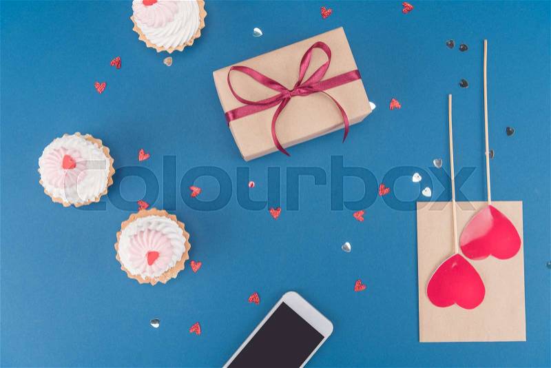 Top view of gift box, smartphone, cakes and envelope isolated on blue, birthday party concept, stock photo