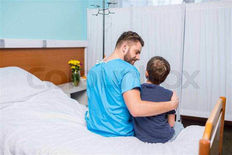Back view of smiling father and little son sitting together on hospital bed, dad and son in hospital , stock photo