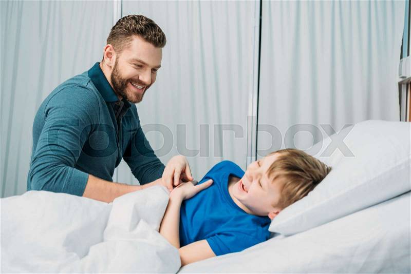 Smiling father playing with sick little boy lying in hospital bed, dad and son in hospital , stock photo