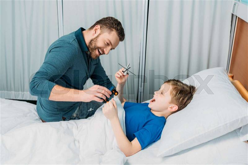 Smiling father playing with sick little boy lying in hospital bed, dad and son in hospital , stock photo