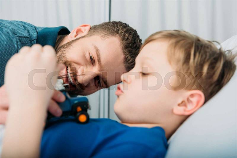 Smiling father playing with sick little boy lying in hospital bed, dad and son in hospital, stock photo