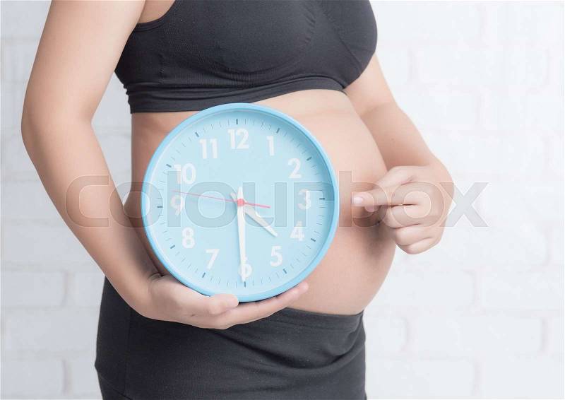 Pregnant woman holding a big round clock against white brick wall, stock photo