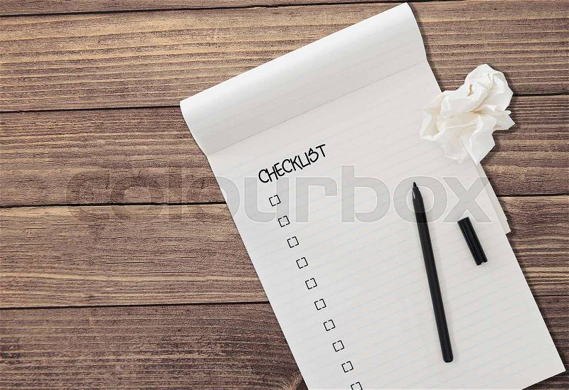 Ruled notepad with word CHECKLIST and check boxes with black felt tip pen and a piece of crumpled paper isolated on rustic wooden table background, stock photo
