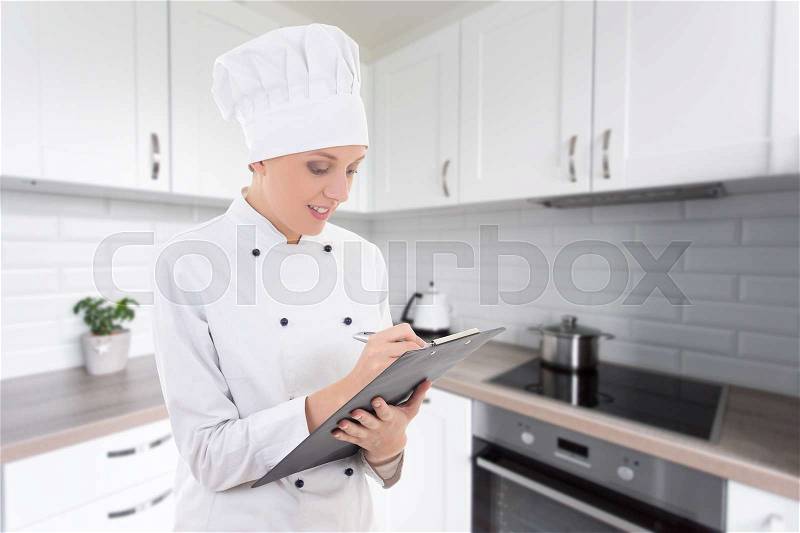 Portrait of young woman in chef uniform writing something in clipboard in modern kitchen, stock photo