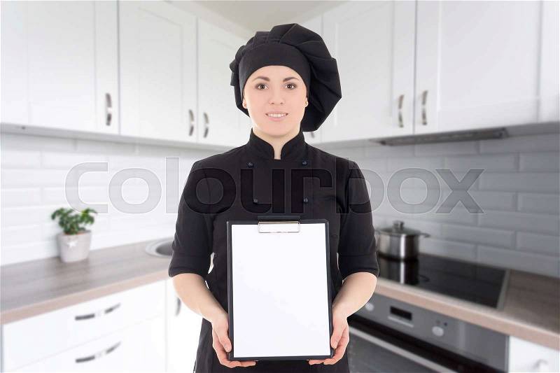 Young cook woman in black uniform showing blank clipboard in modern kitchen, stock photo