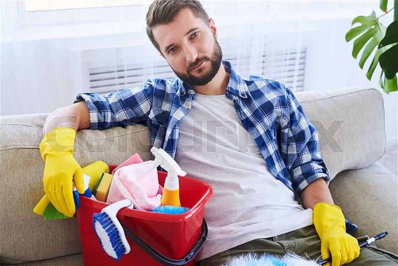 Mid shot of tired guy sitting on sofa with cleaning set, stock photo