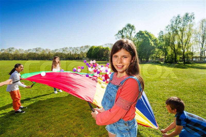 Portrait of girl throwing colorful balls playing rainbow parachute game on the green field in spring, stock photo