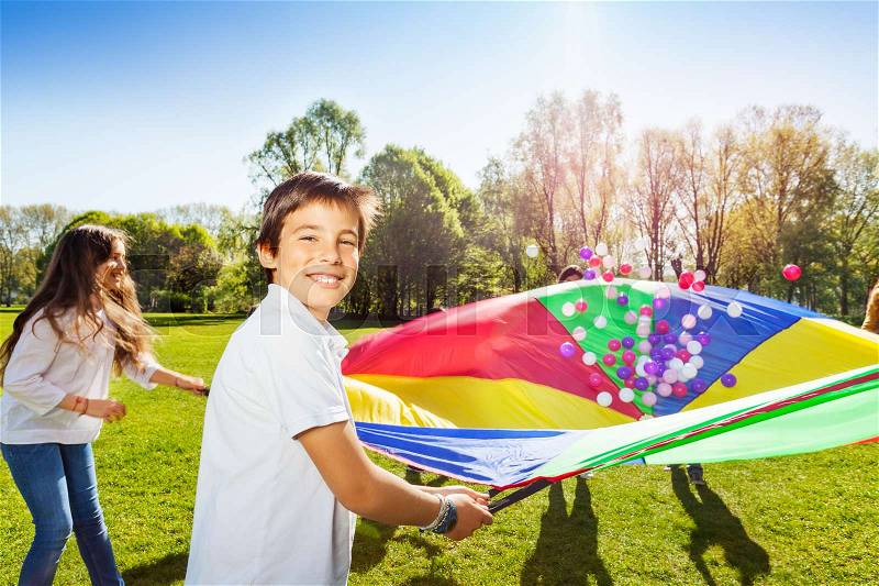 Portrait of happy boy holding rainbow parachute full of colorful balls and playing with friends in the park, stock photo