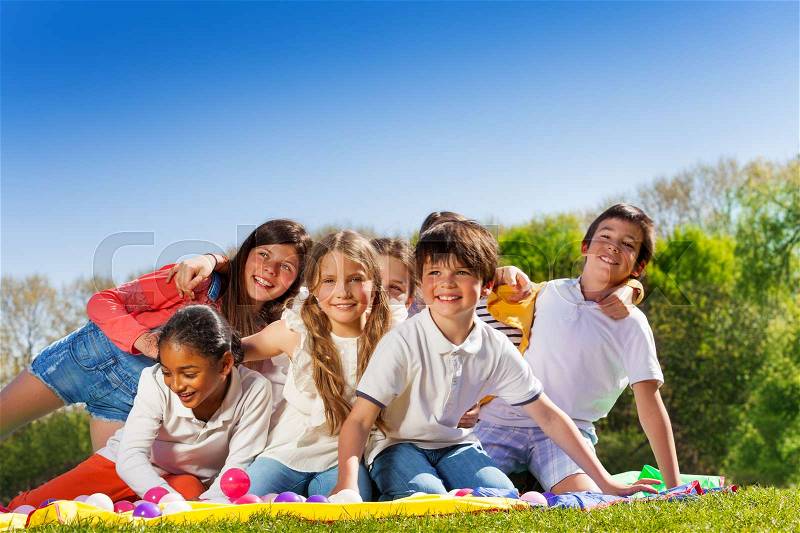 Big group of happy 10-12 years old kids, sitting on the lawn in the park, hugging and laughing, stock photo