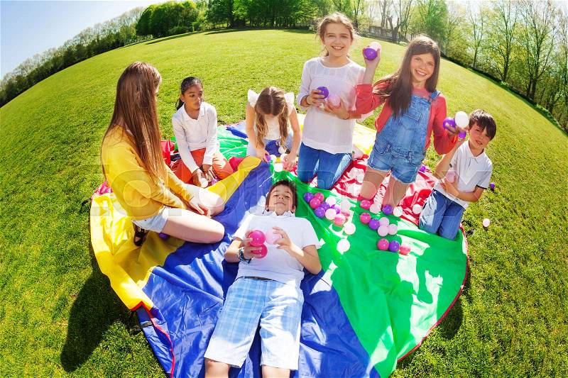 Big group of happy kids sitting or laying on rainbow parachute and playing with colorful balls in the park, stock photo