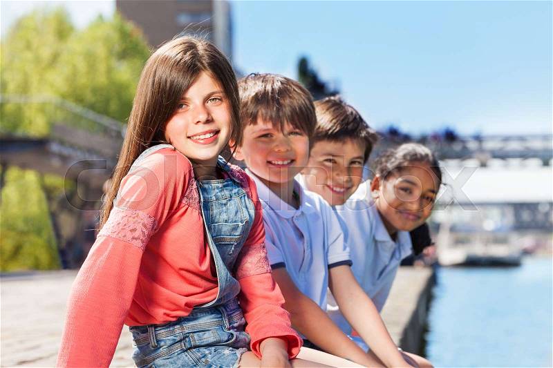 Close-up portrait of happy schoolchildren sitting in a line on bank of river in summertime, stock photo