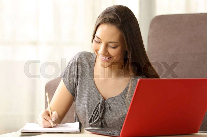 Student learning and taking notes in a paper notebook with a red laptop beside sitting on the floor in the living room at home, stock photo