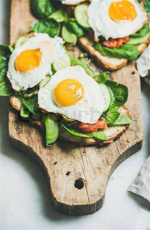 Healthy breakfast sandwiches. Bread toasts with fried eggs and fresh vegetables on rustic wooden board over grey marble background, selective focus. Clean eating, healthy, weight loss food concept, stock photo