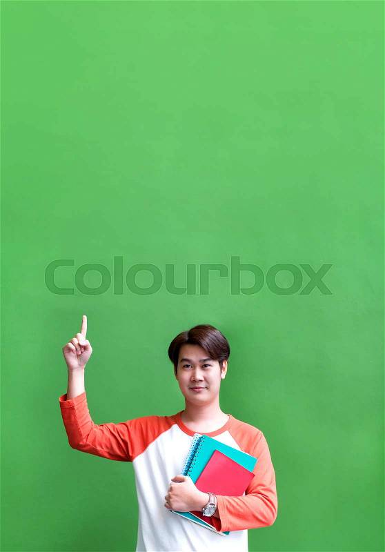 Teenager man point up at copy space over head and holding notebook at green wall in university campus,Copy space for adding content of design for advertising in education campaign concept., stock photo