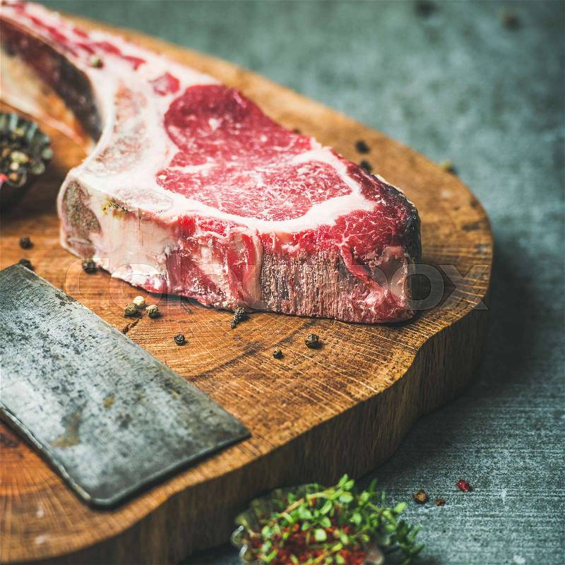 Dry aged raw beef rib eye steak with bone, butcher meat chopping knife and spices on wooden board over grey concrete background, selective focus, stock photo