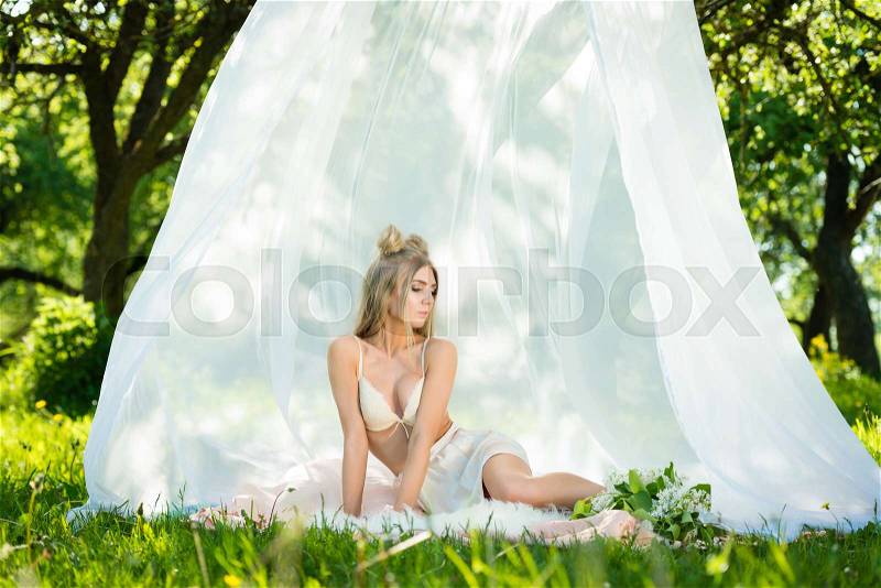Sexy young woman in bra and mini skirt sitting in tent with lilac flowers, stock photo