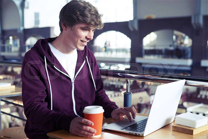 Mid shot of smiling guy printing in laptop over paper cup of coffee while sitting in café, stock photo