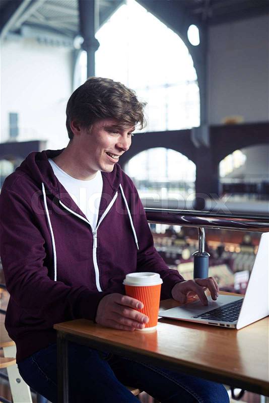 Vertical of delightful man printing in laptop over paper cup of coffee in cafe, stock photo