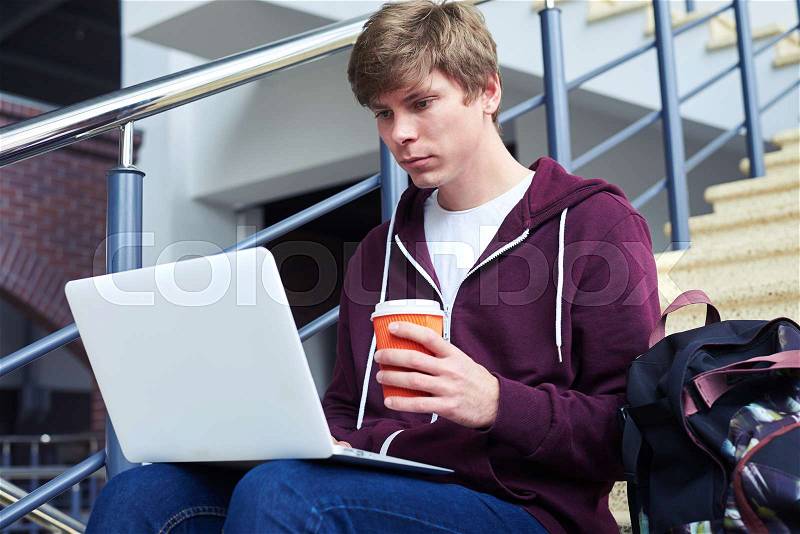 Mid shot of boy with poker face holding cup of coffee while surfing in laptop, stock photo