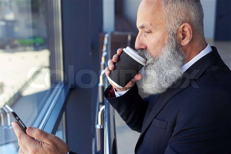 Close-up of serious old gentleman drinking coffee and surfing in phone while leaning on handrail, stock photo