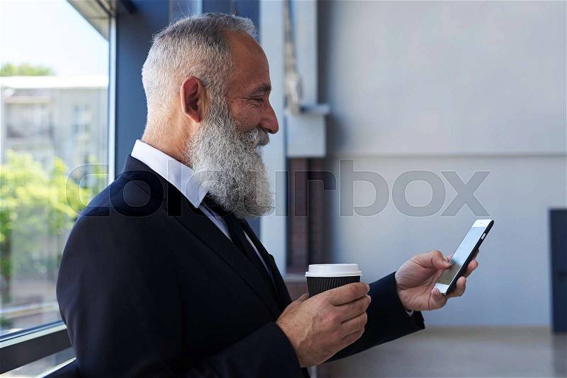 Close-up of smiling old man drinking coffee and surfing in phone while leaning on handrail, stock photo