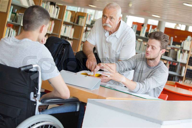 Student in wheelchair talking with classmate and teacher in library, stock photo