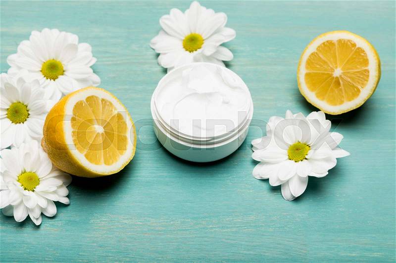 Close-up view of organic cream in container, sliced lemon and chamomiles on blue wooden table, stock photo