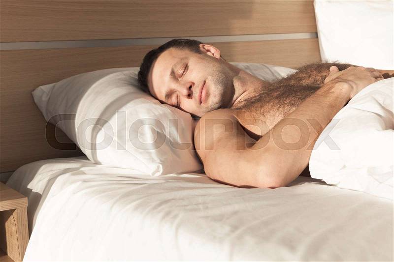 Young European man sleeping in wide bed under white blanket in bright morning sunlight, stock photo
