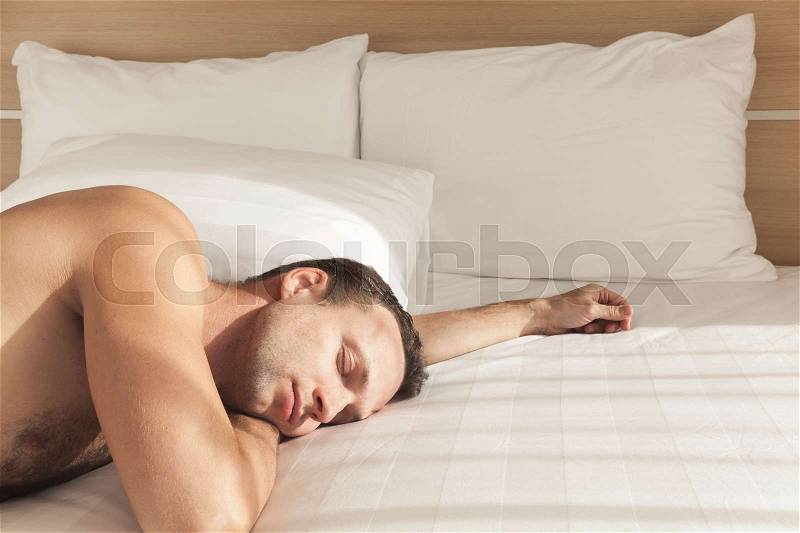 Young European man sleeping in wide bed in morning sunlight, stock photo
