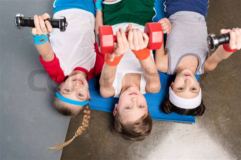 Top view of cute children in sportswear lying on yoga mat and exercising with dumbbells in gym, children sport concept, stock photo