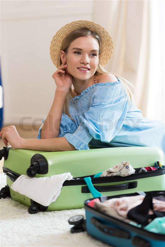 Smiling young blonde woman in hat packing suitcase and looking away, packing luggage concept, stock photo