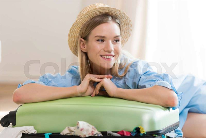 Smiling young blonde woman in hat packing suitcase and looking away, packing luggage concept, stock photo
