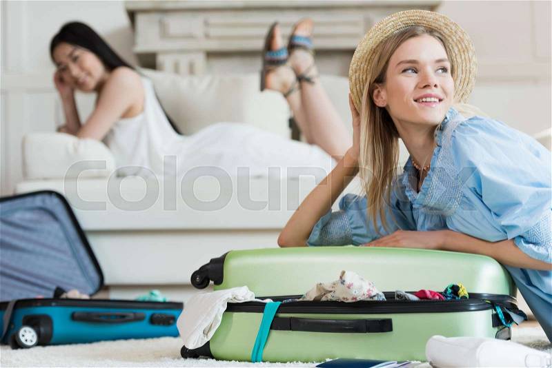 Smiling young woman in straw hat packing suitcase and looking away, packing luggage concept, stock photo