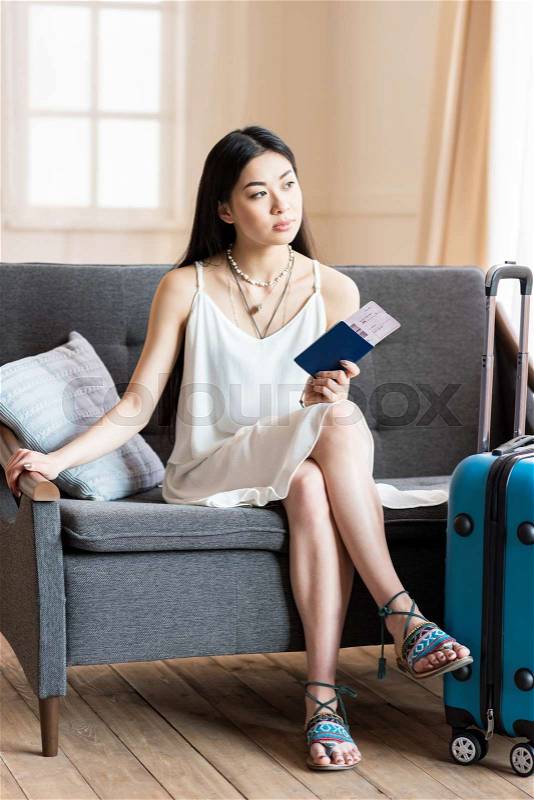 Asian woman traveler sitting on sofa with suitcase and passport, packing luggage, stock photo