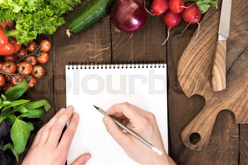 Food composition of raw vegetables, chopping board and human hand able to write something in notepad on wooden background , stock photo